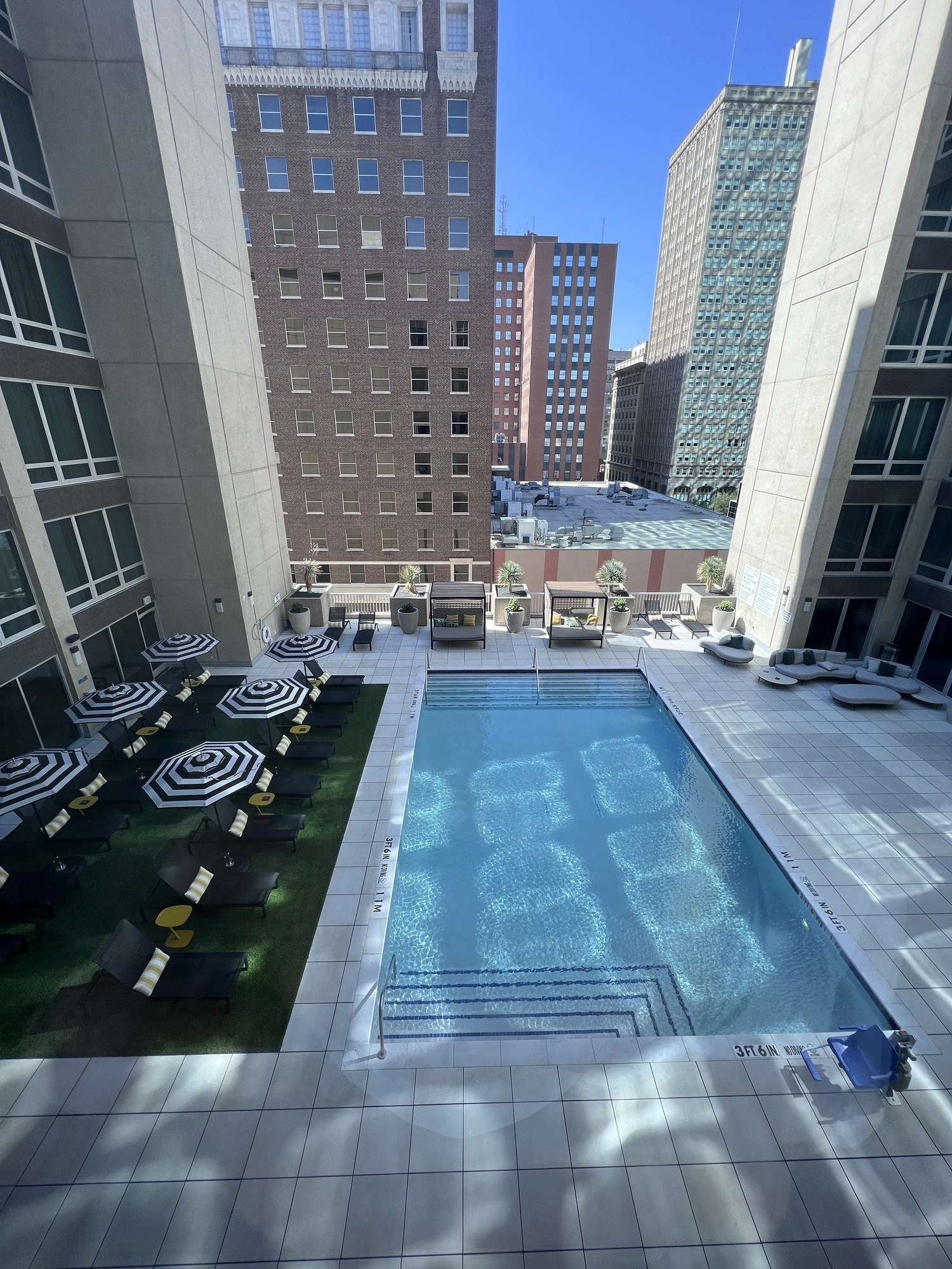 A pool seen from above in a courtyard between brick and glass towers, with striped parasols and loungers along one side