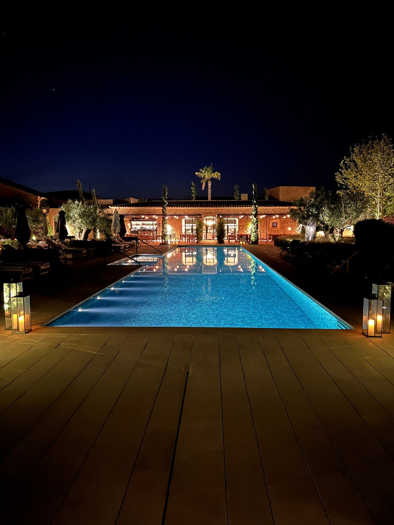 A long pool lit at dusk on a timber deck, the lit house and palms reflected in it