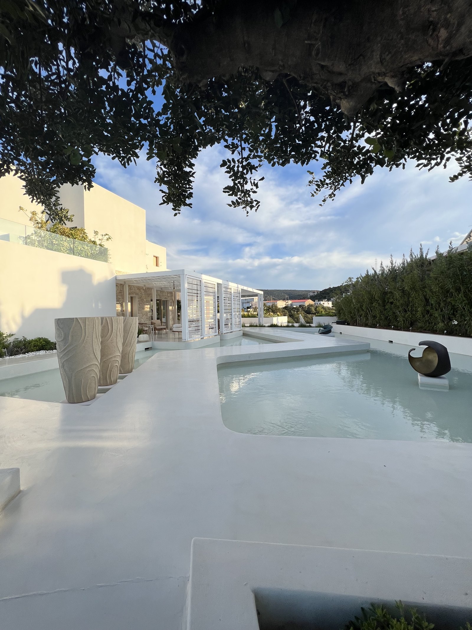 A white pool and terrace seen from under the dark canopy of a tree, with ribbed white stools along the edge and the hills behind