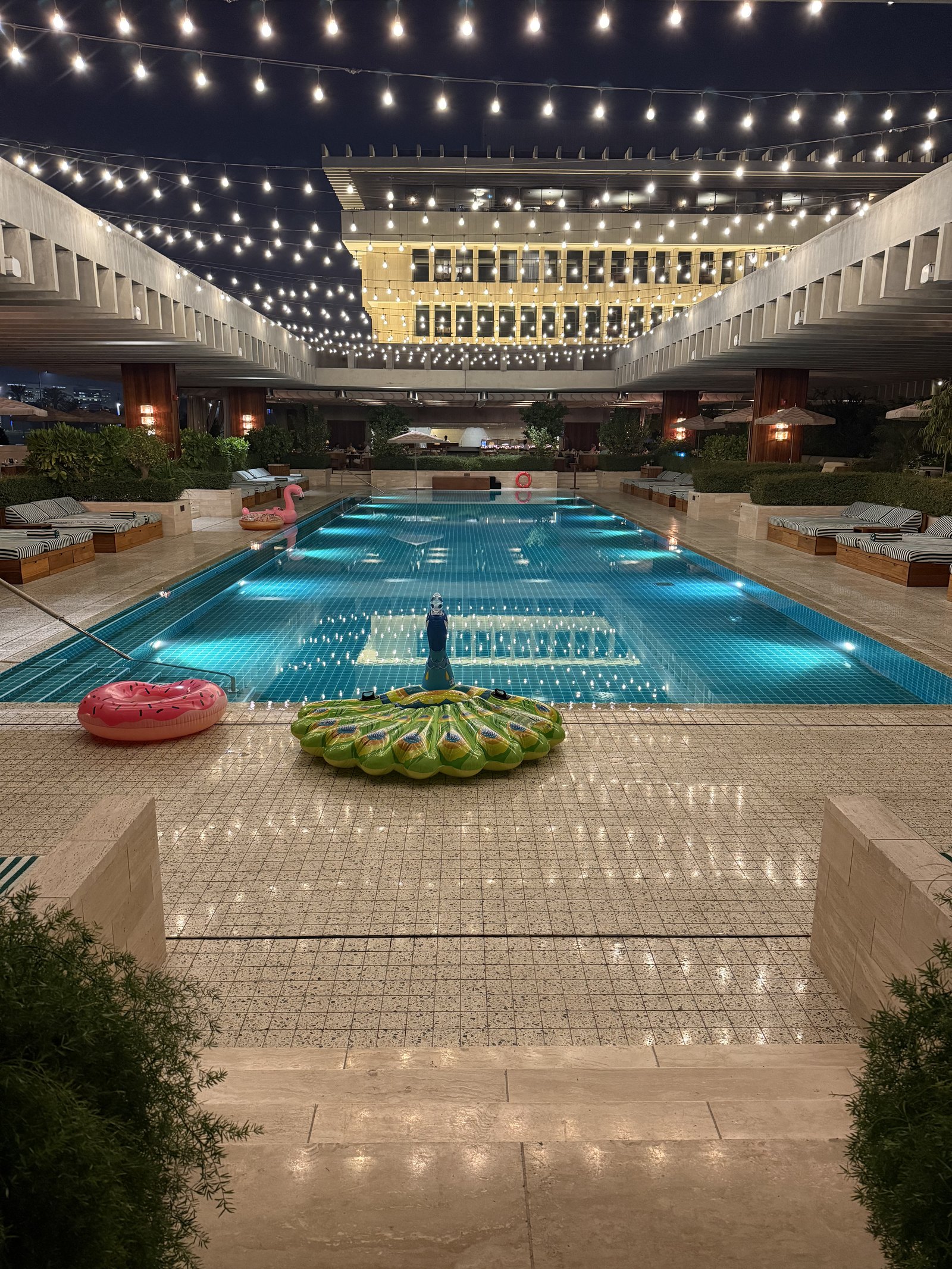 A long pool lit blue at dusk between colonnaded wings, under strings of festoon lights