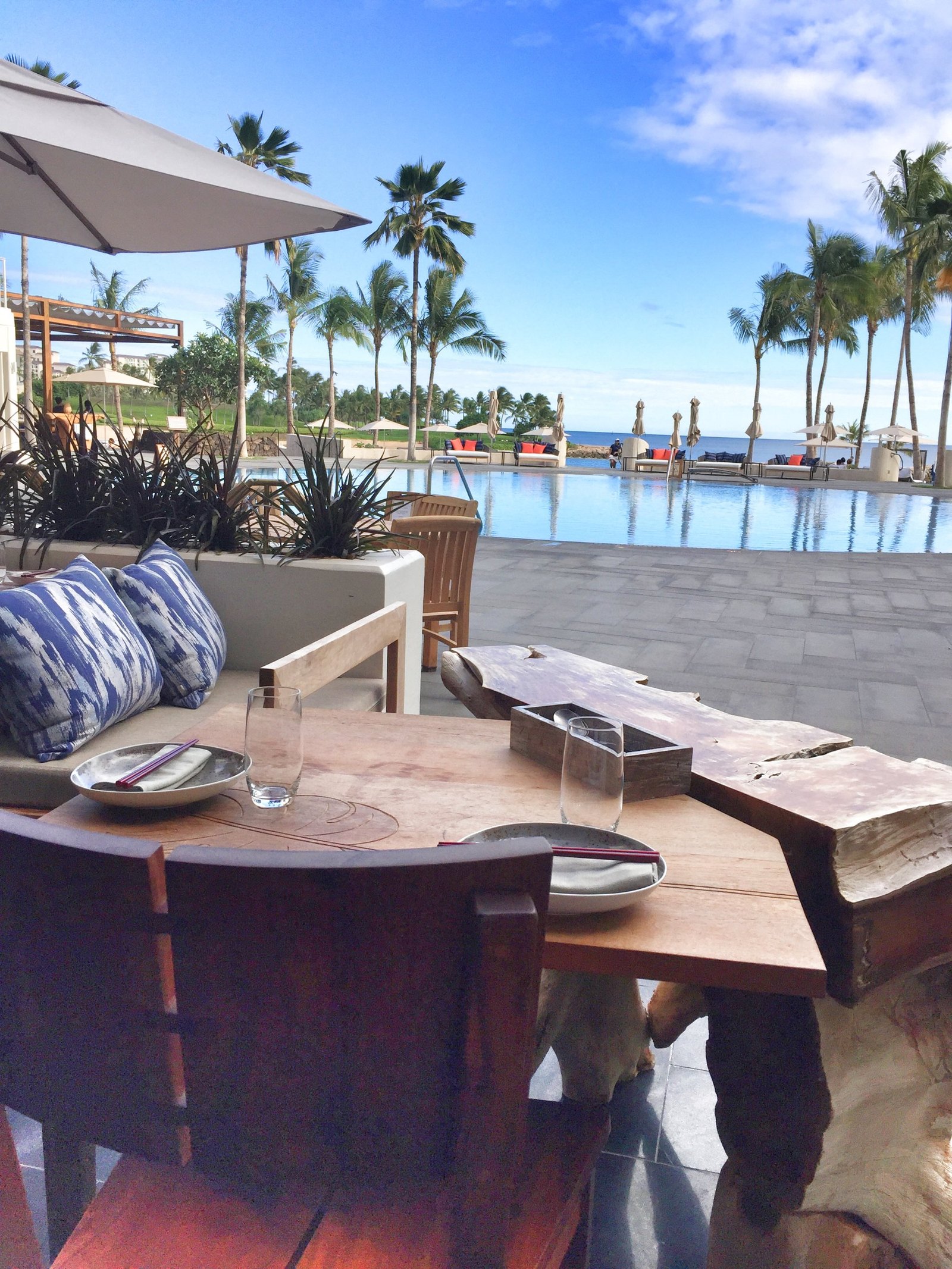A restaurant table of live edge timber set with chopsticks on a terrace above an infinity pool, palms and the sea beyond