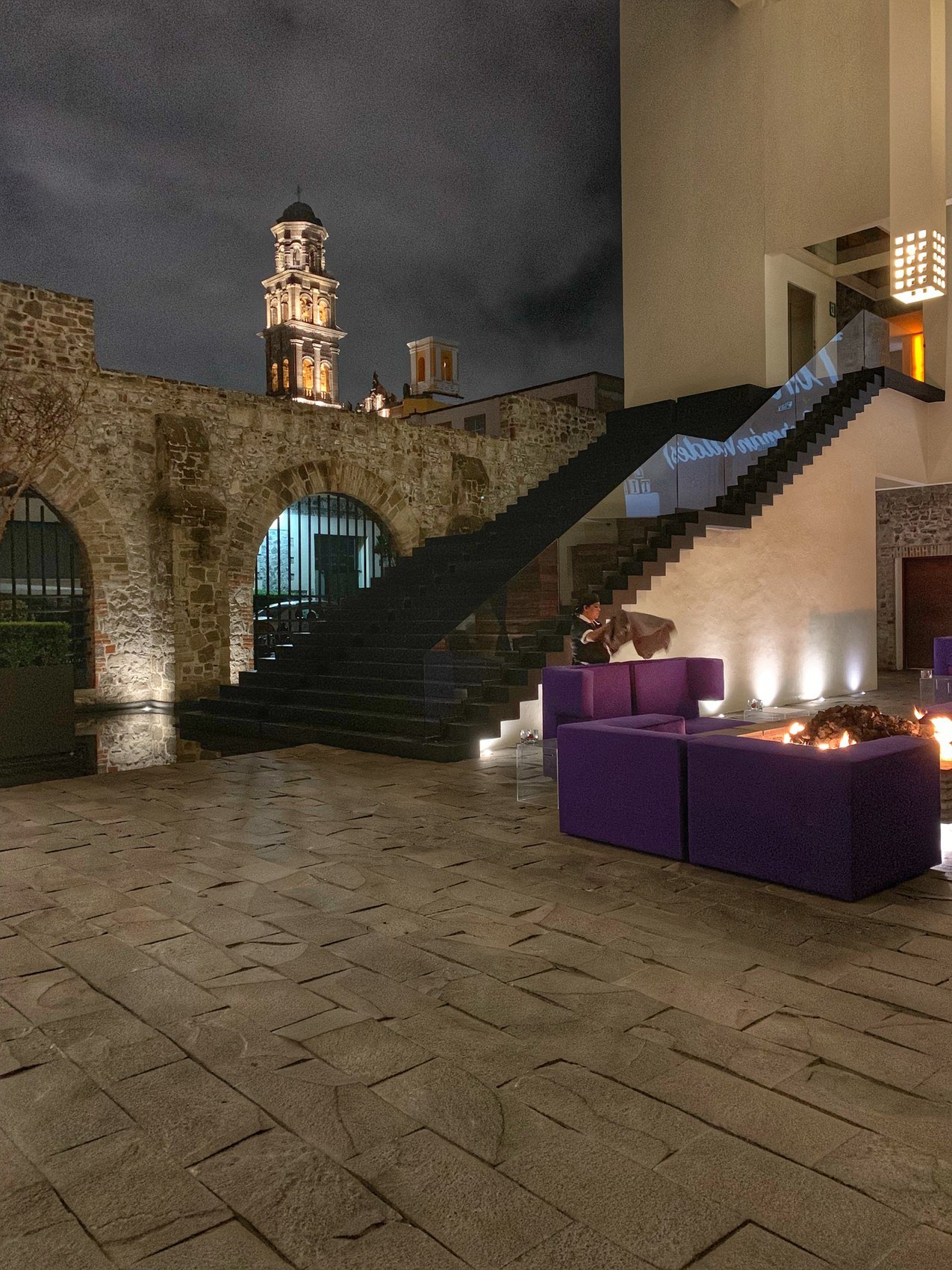 A black staircase rising beside a lit stone arch, a church tower beyond it