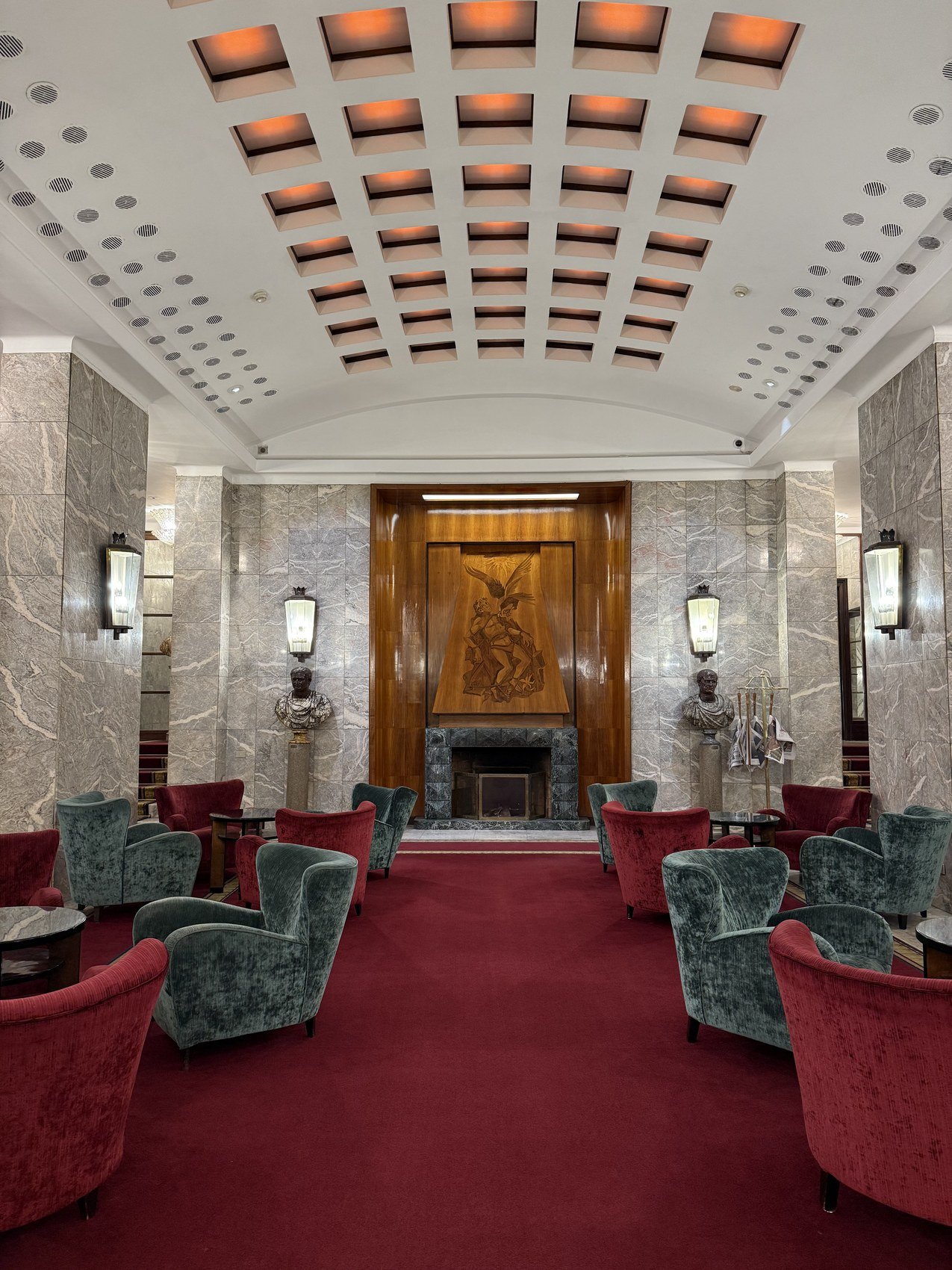 A lobby of grey marble and backlit coffering with pale blue armchairs on a red carpet