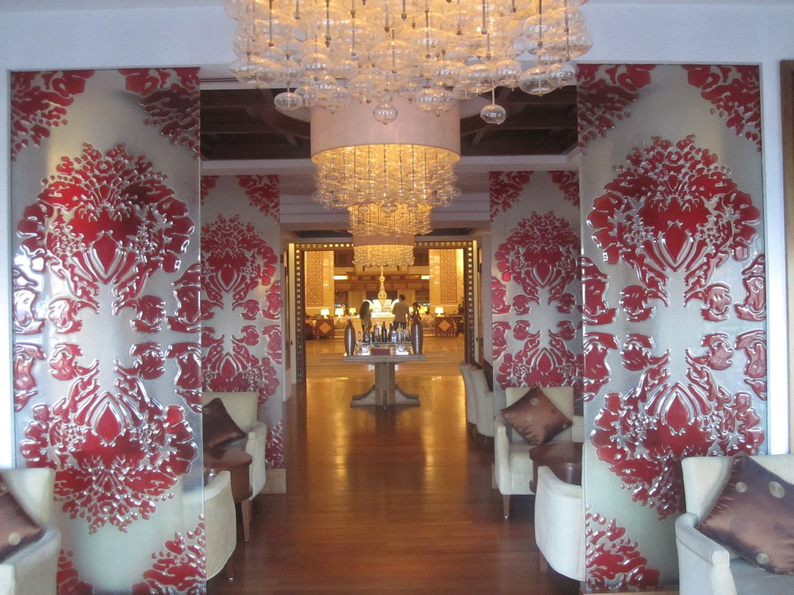 A lobby seen between glass panels printed with red damask, under tiers of clear bubble glass chandeliers