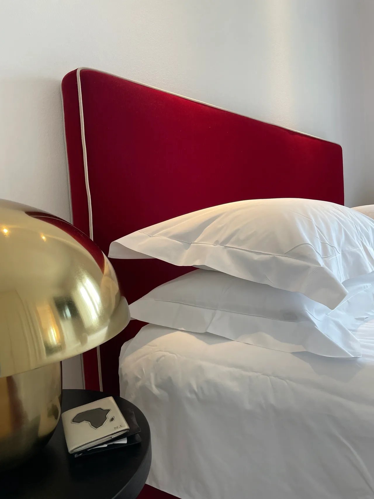 A bed with a red velvet headboard piped in white and white pillows stacked against it, beside a domed brass lamp on a black table with a small notebook under it