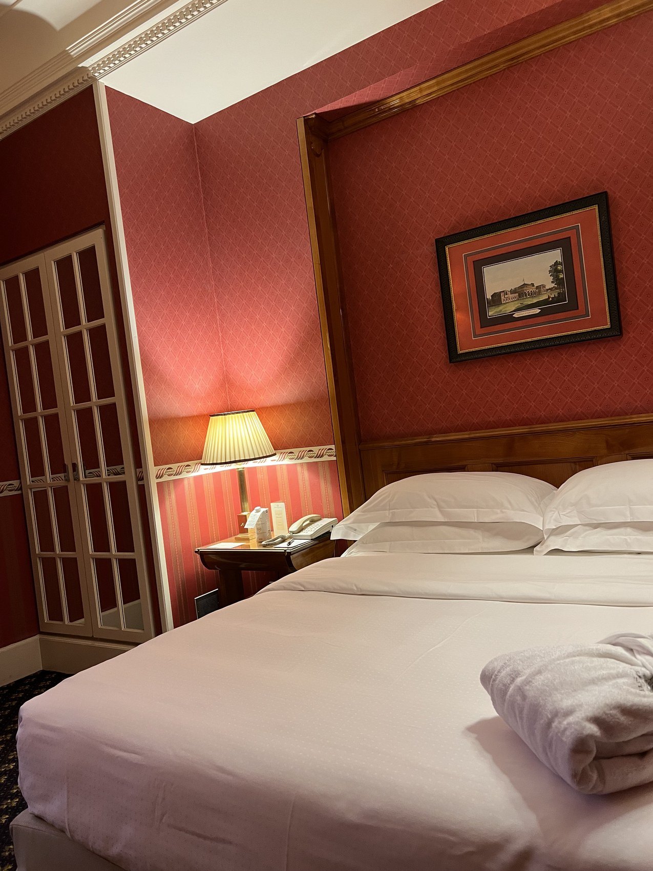 A bed against a red padded wall with a brass framed door and a small framed print