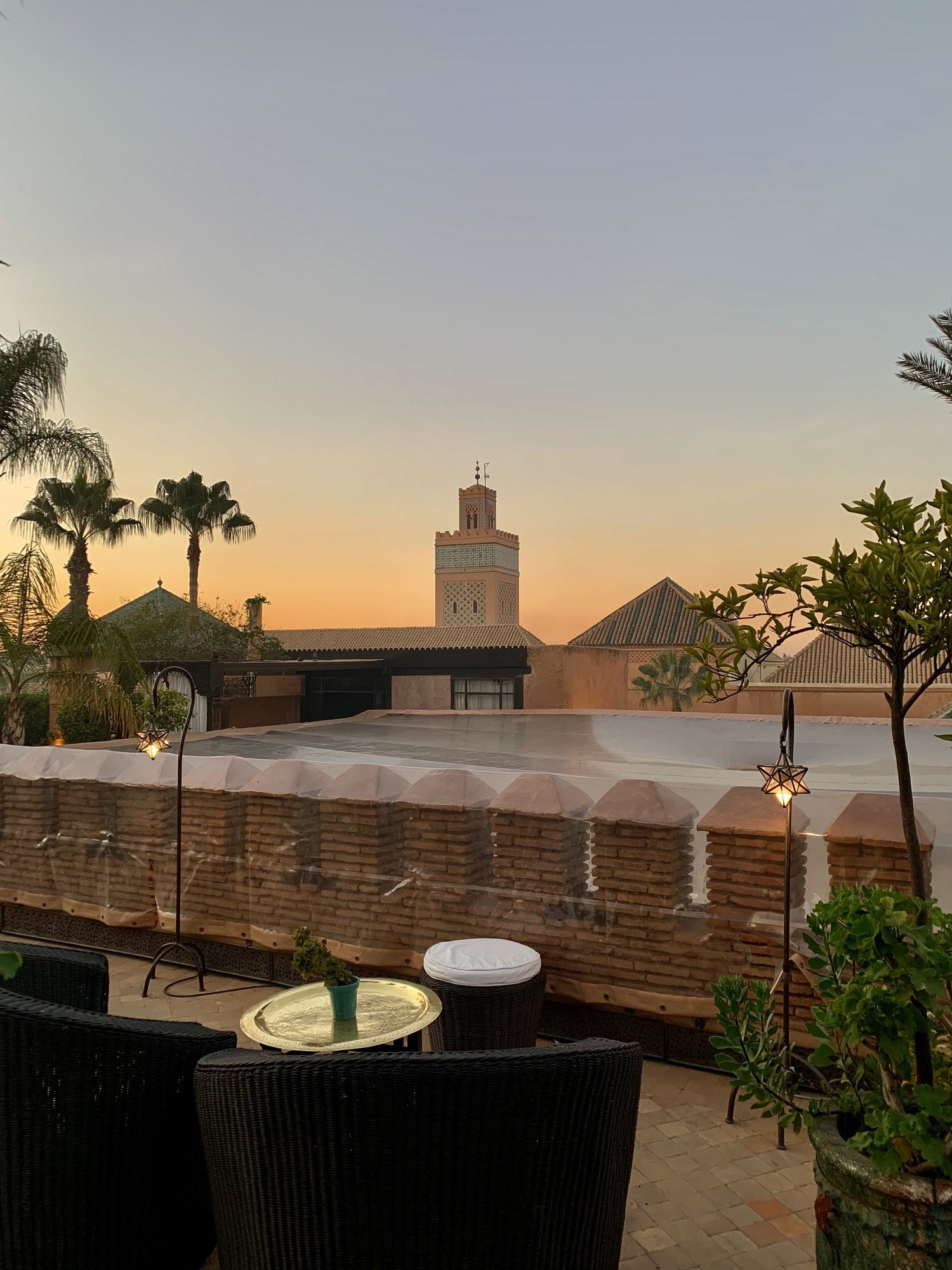 A low seating area with dark armchairs on a roof terrace at dusk, looking across the flat roofs of the medina to the Koutoubia minaret