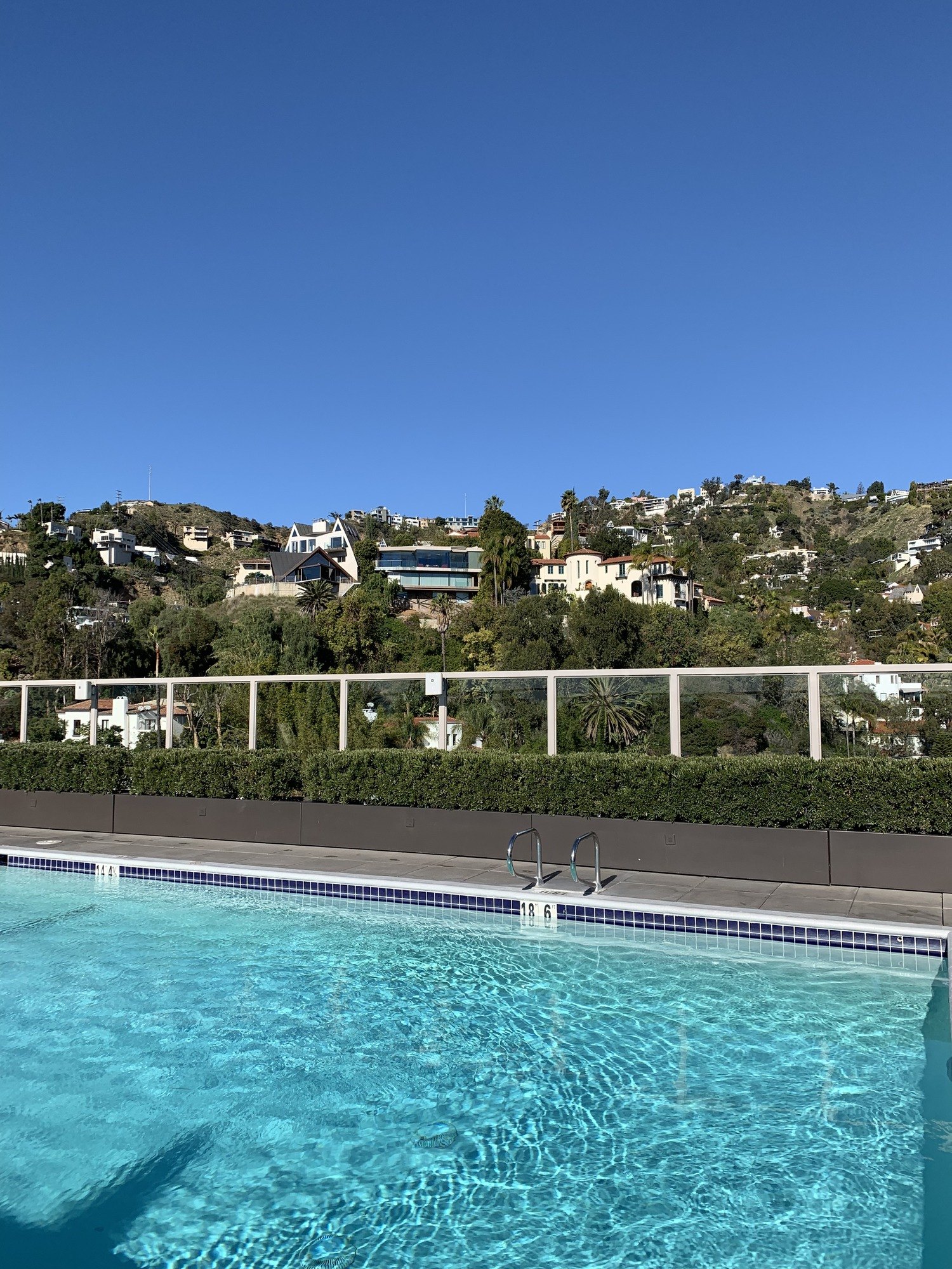 A rooftop pool with the water lit turquoise, looking across a glass rail to wooded hills and scattered houses under a clear sky