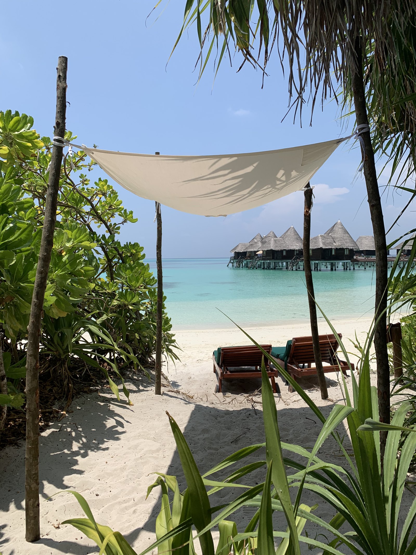 A white sail canopy strung between poles over two loungers on the sand, with overwater villas across the lagoon