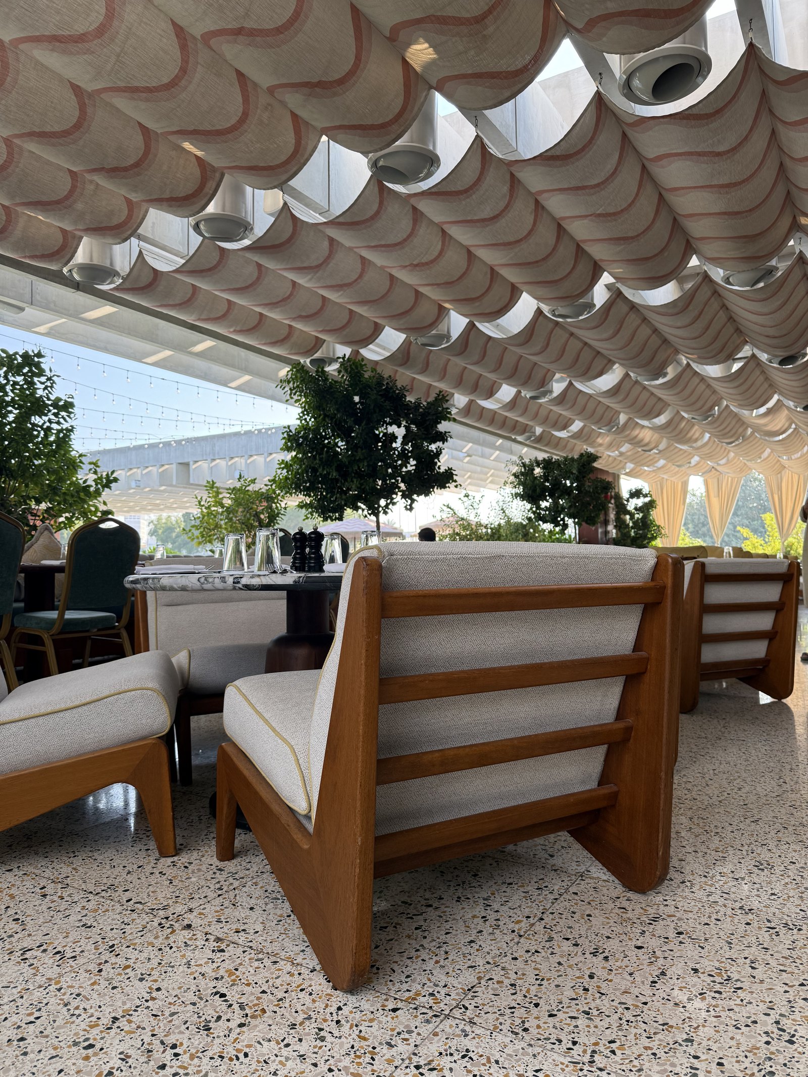 A terrace restaurant under a canopy of overlapping pale scallops, with deep slatted armchairs, potted trees and a terrazzo floor
