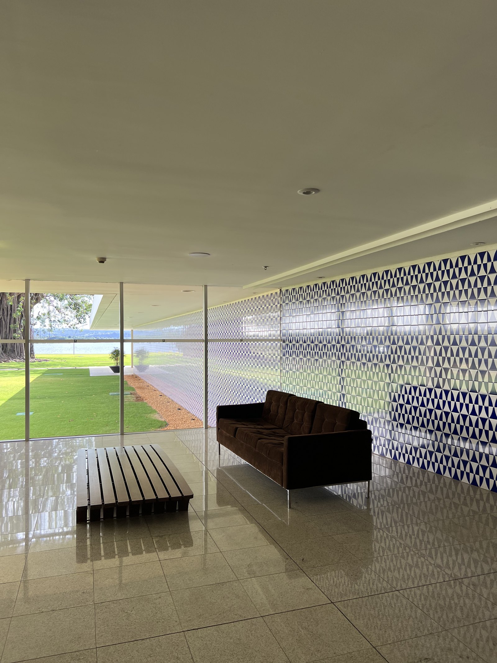 A long room with a patterned perforated screen down one side, a dark leather sofa and a slatted bench on a polished floor, open through full-height glass to a lawn