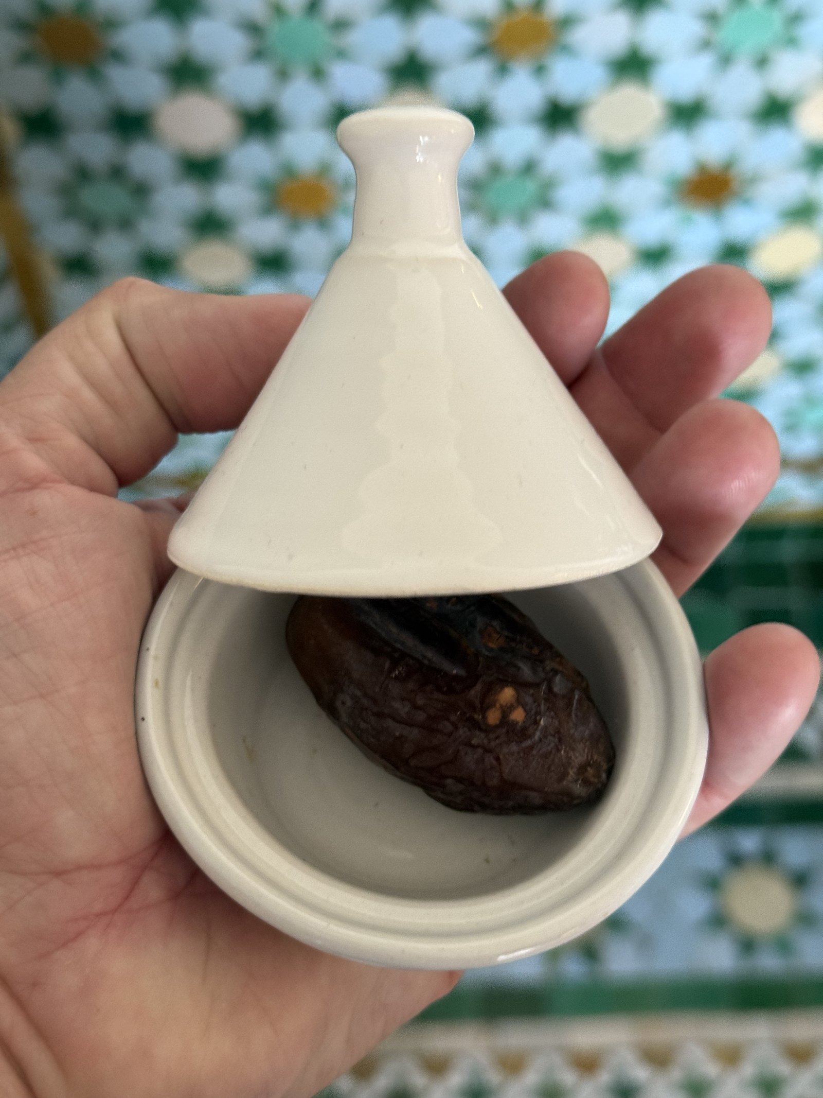 A small white ceramic tagine held in one hand with the lid lifted and a date inside it, against a wall of green, white and ochre star-patterned zellige