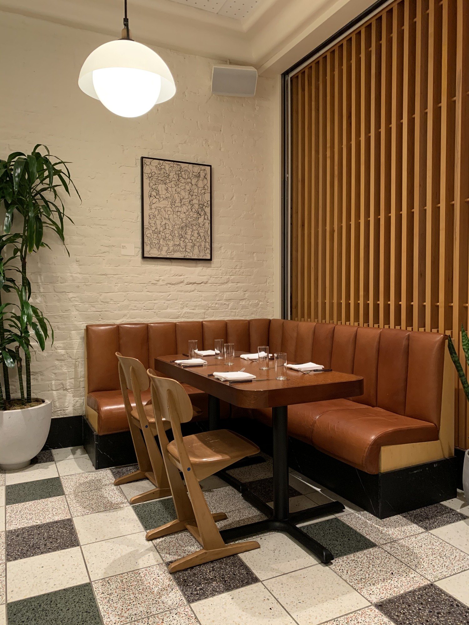 A tan leather banquette and pale timber chairs at a laid table against a whitewashed brick wall, with a framed drawing above and a tall plant beside