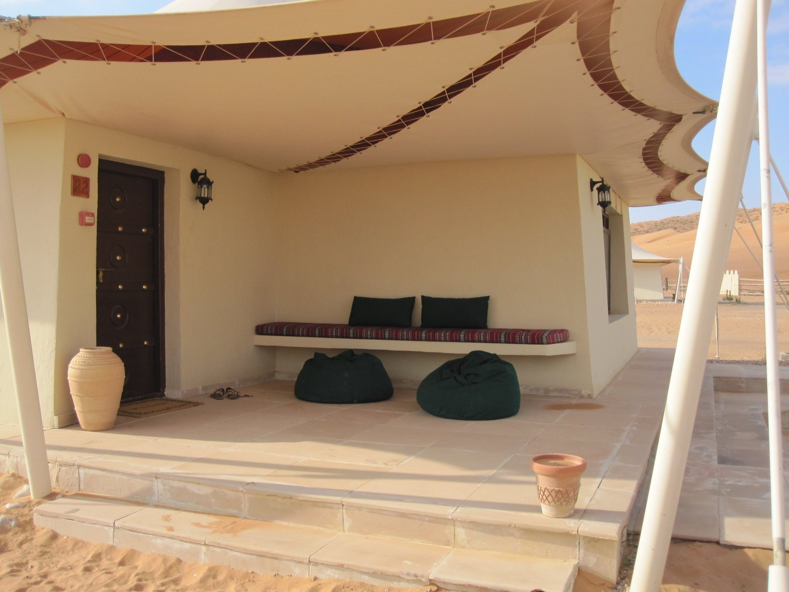 The shaded porch of a tent with a cushioned bench, two dark bean bags, a terracotta jar beside the door and dunes beyond