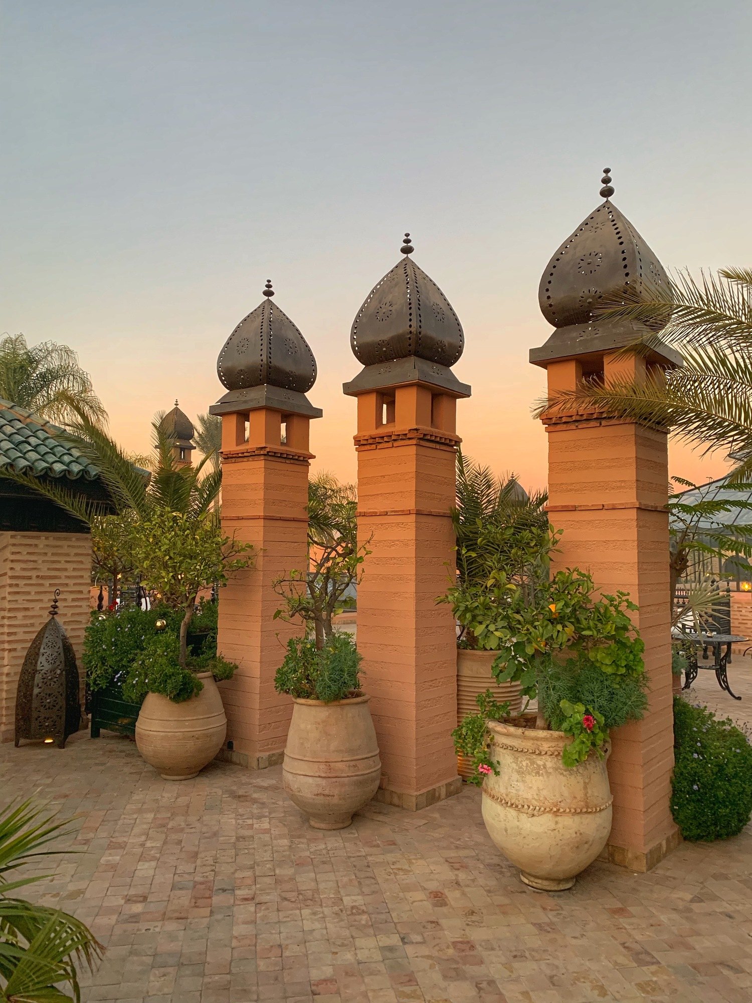 Three rendered towers with pointed caps standing above a line of terracotta urns planted with palms, against a pale dusk sky