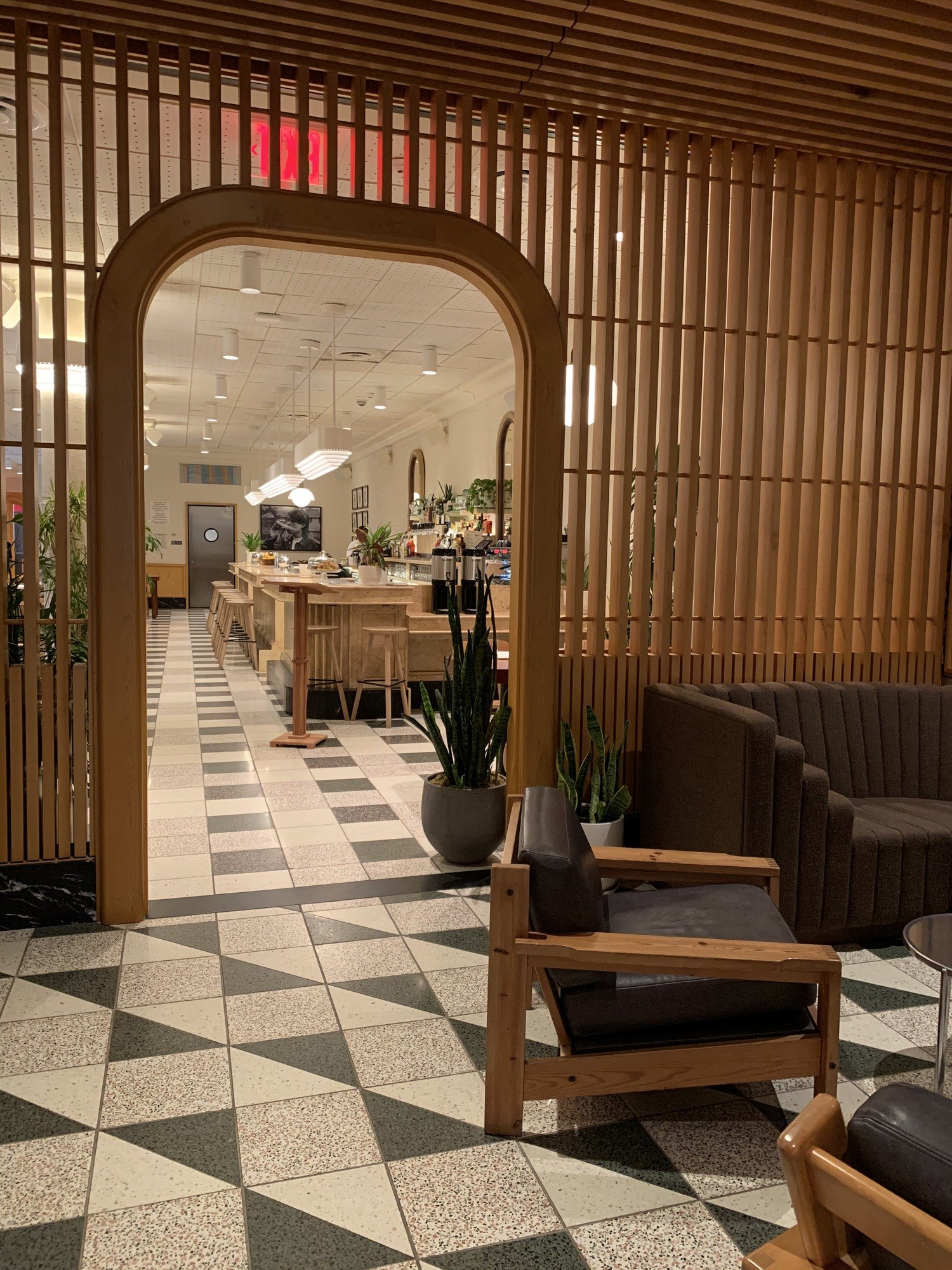 A long room seen through an arch in an oak screen, with the checkerboard floor running away to a lit bar, leather and timber chairs and planted pots along the way