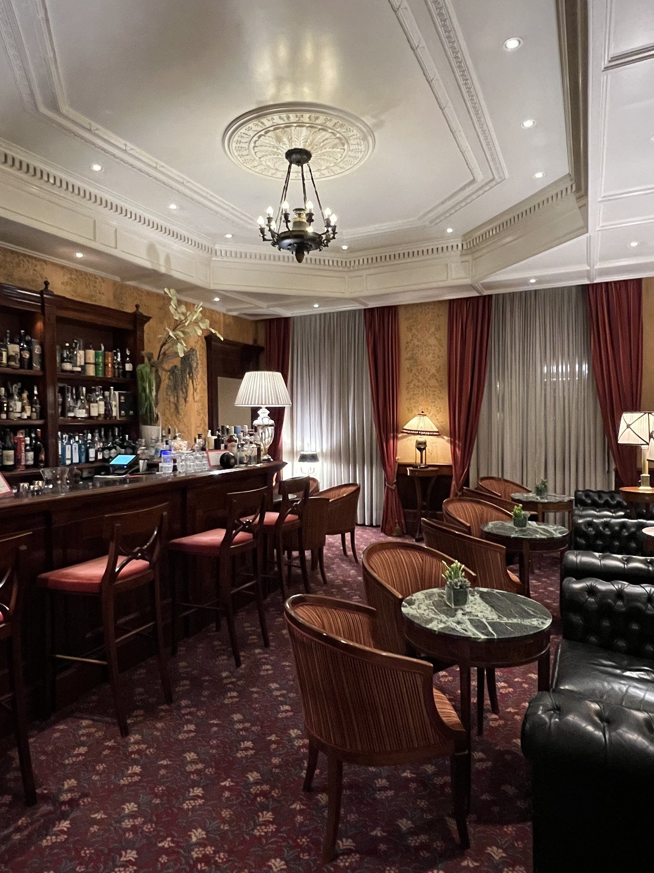 A timber bar with a chandelier above, cane chairs and red velvet curtains