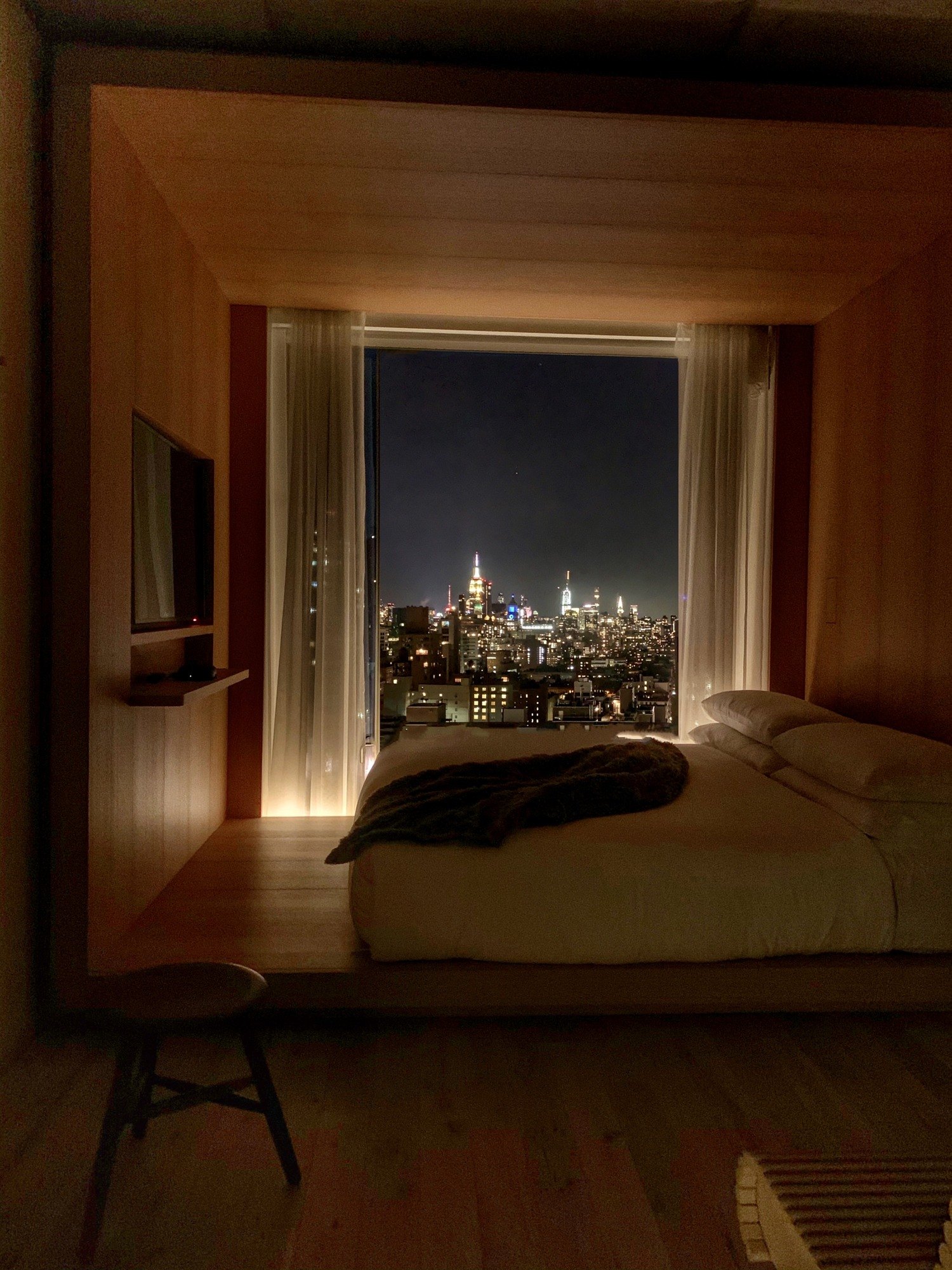 A bed in a room lined entirely in pale timber, looking out through a deep window to the lit towers of the city at night