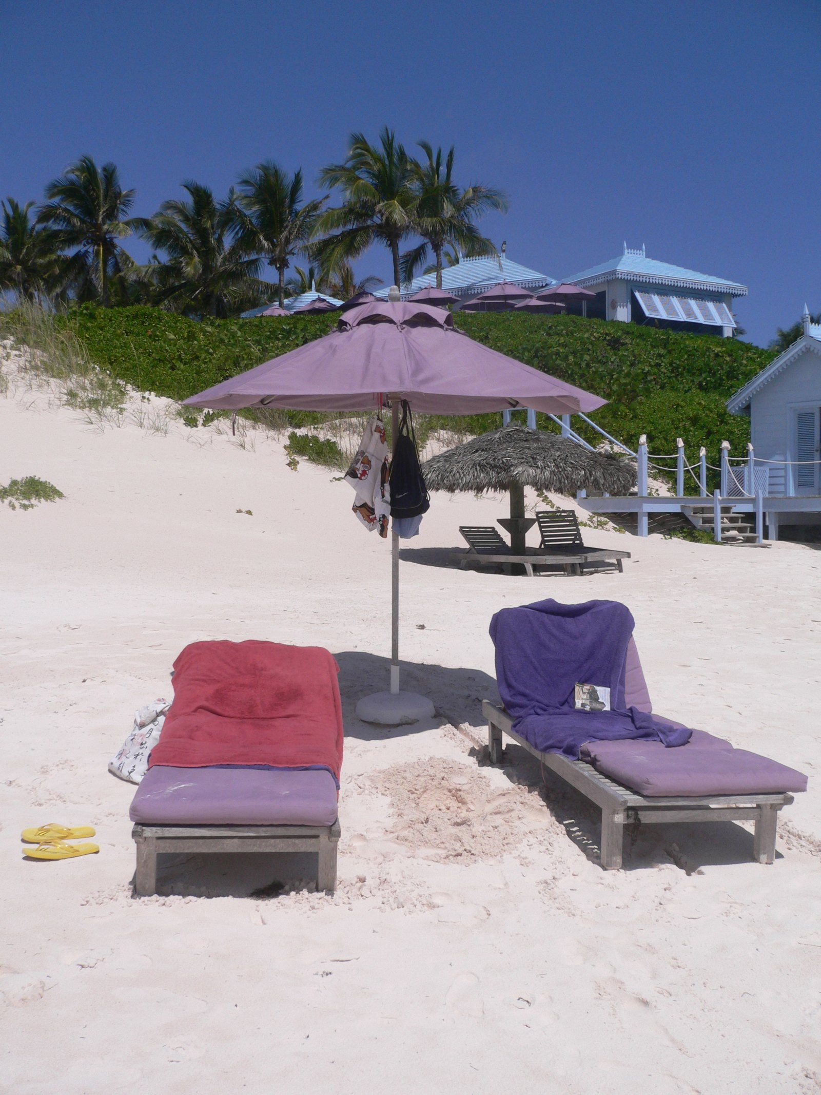 Two loungers dressed in red and purple towels under a lilac parasol, pale blue cottages on the dune above