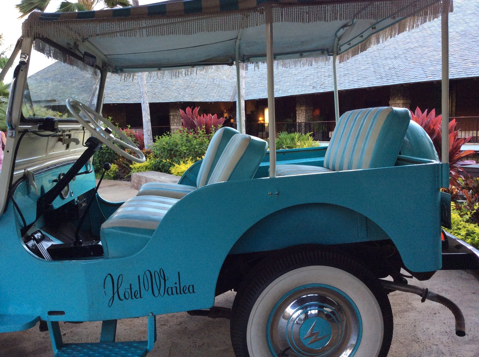 A turquoise open jeep lettered Hotel Wailea, with striped seats and a fringed canvas canopy, parked in front of a shingled building