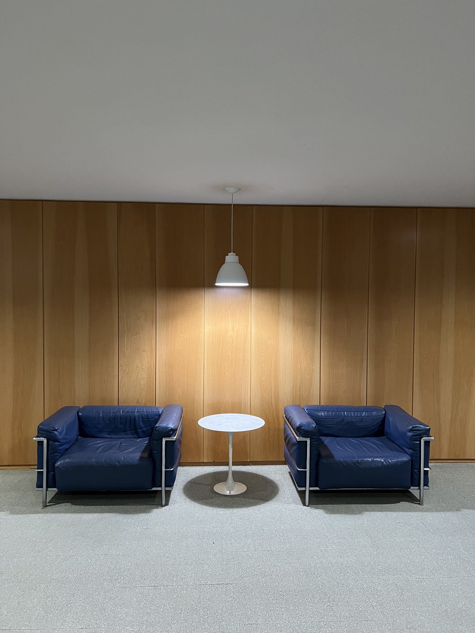 Two blue leather armchairs on chrome frames either side of a small round table, against a wall of pale timber panelling, under one pendant lamp