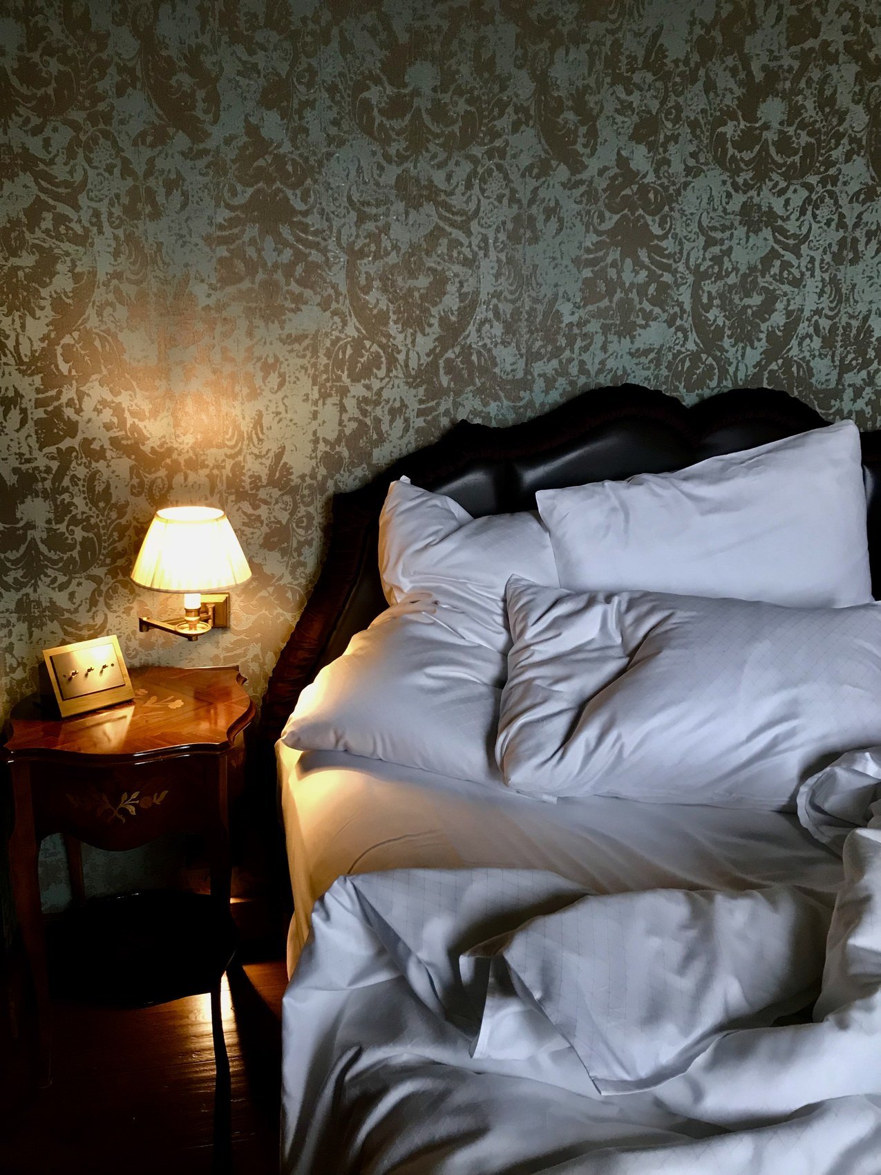 An unmade bed with a carved dark headboard against gold and green damask, a small lamp lit beside it