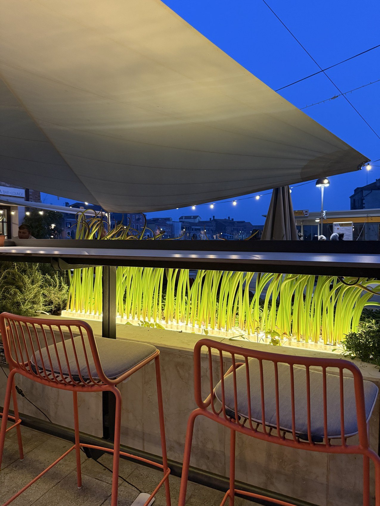 Red metal stools at a rooftop counter at dusk, a lit planter of grasses and a sail awning overhead