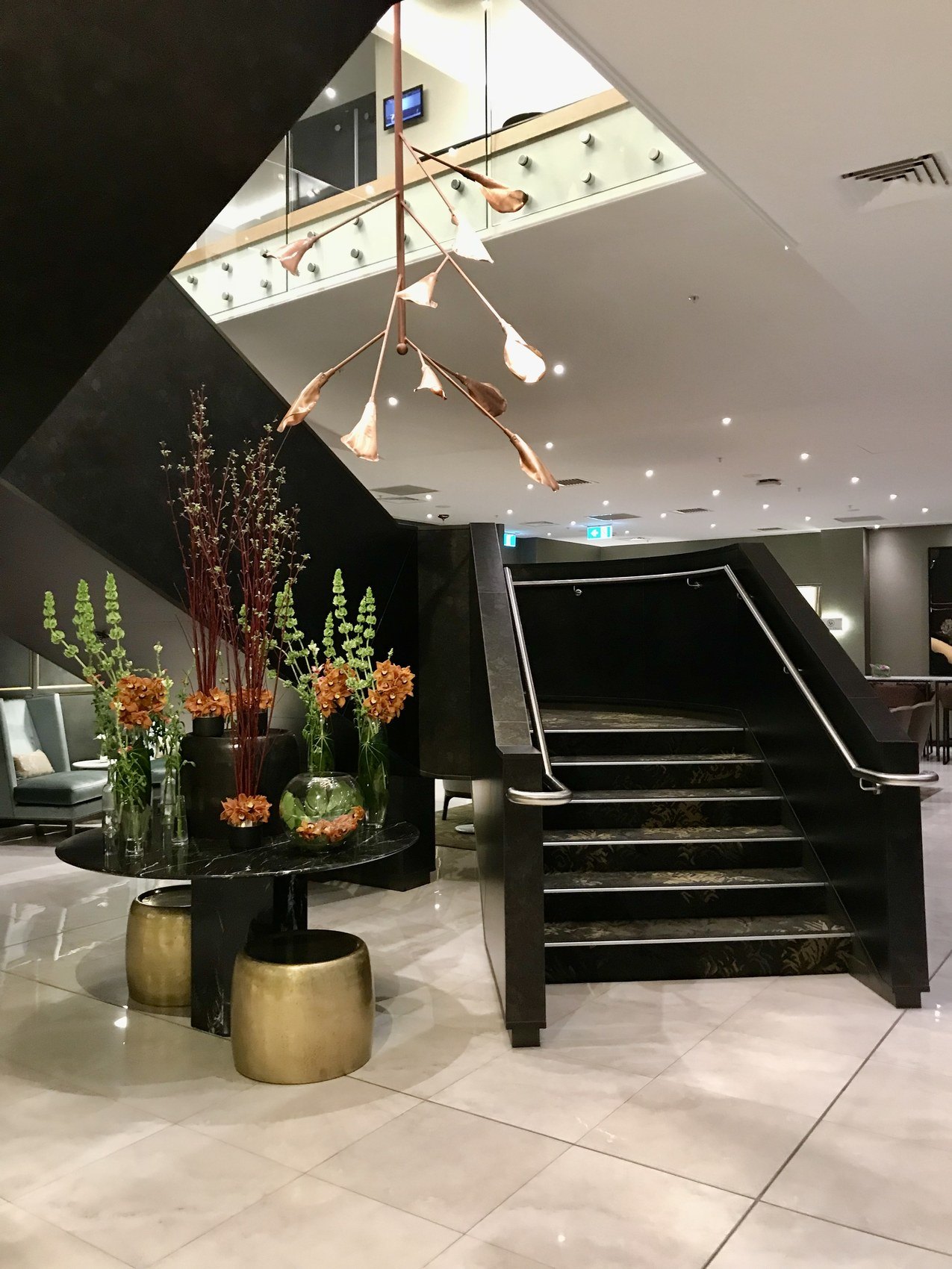 A black staircase in a pale lobby, flowers on brass tables at its foot