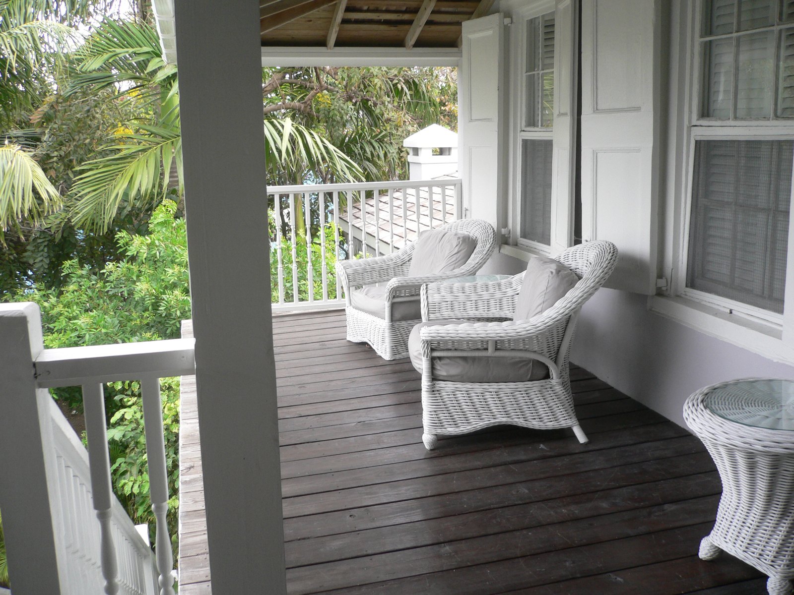 White wicker armchairs on a shaded timber veranda with louvred shutters and palms beyond the rail