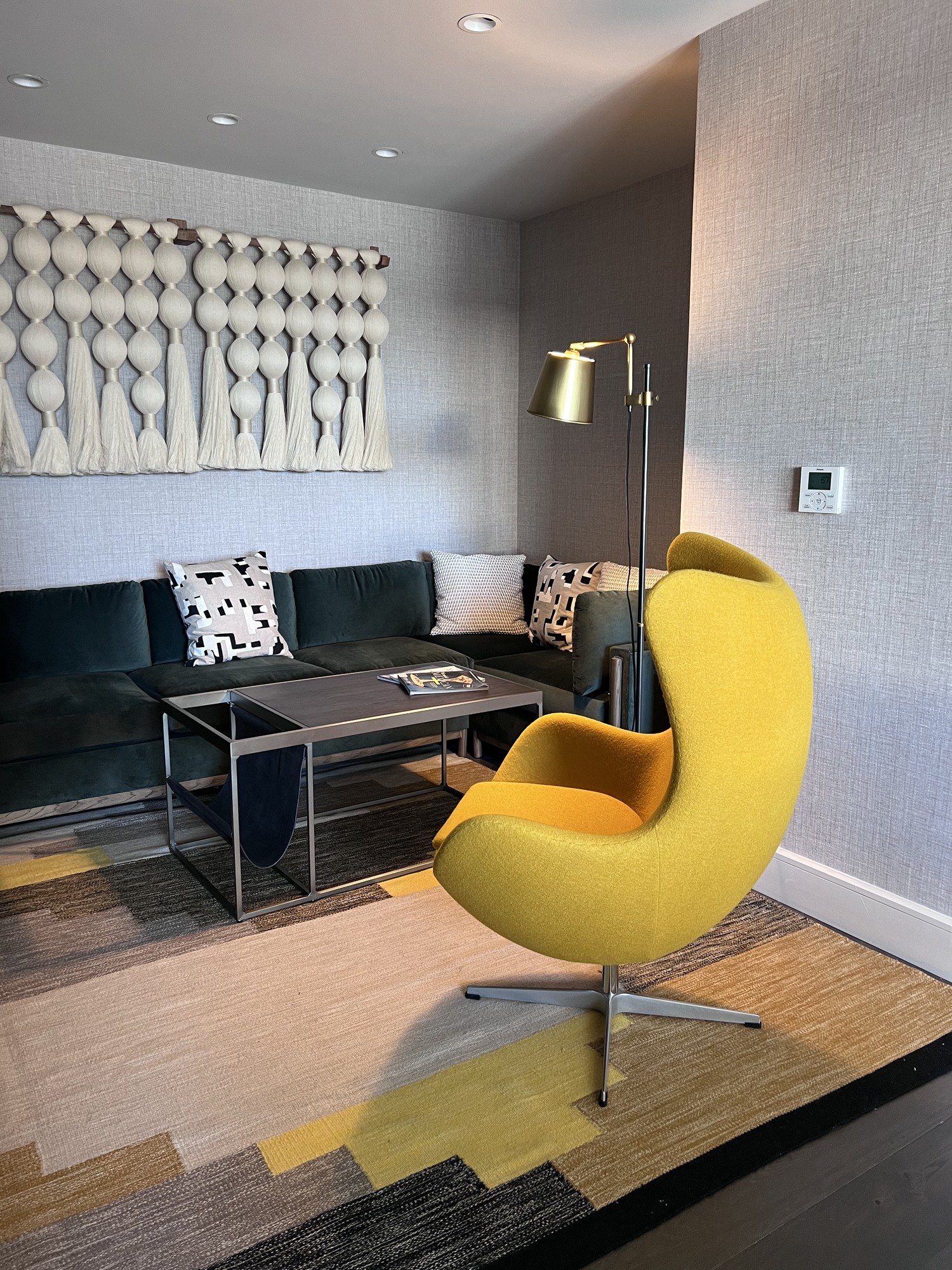 A sitting area in a suite with a yellow swivel armchair, a dark sofa under a woven hanging of pale diamonds, a brass floor lamp and a striped rug