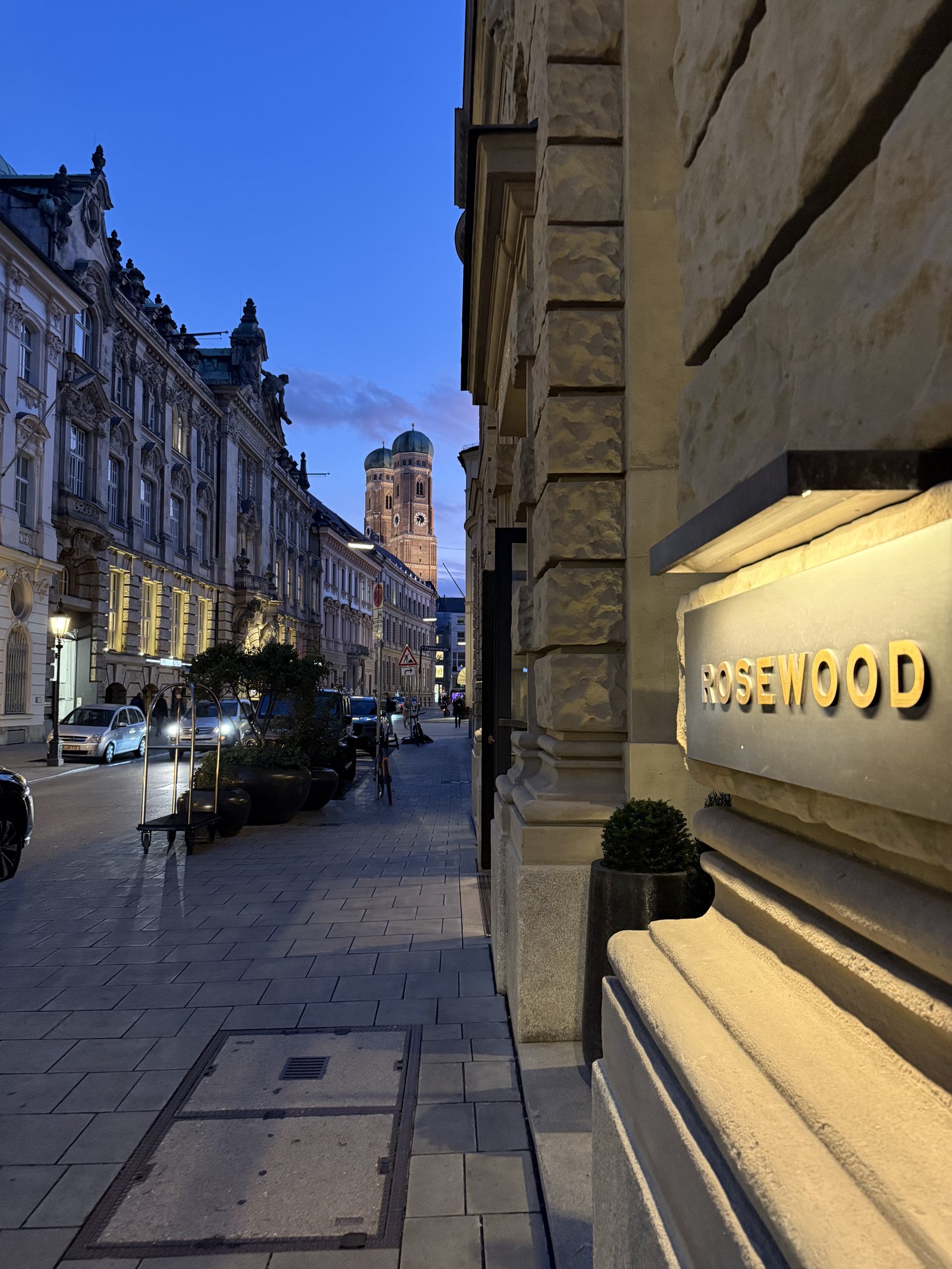 The brass lettering at Rosewood Munich at dusk, with the towers of the Frauenkirche down the street
